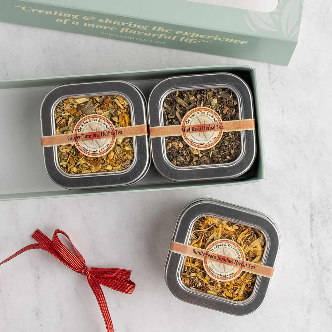 Two square metal tins of loose-leaf herbal tea displayed in a mint green gift box, with a second tin placed outside the box on a light marble surface. The tins have silver lids, round beige labels reading 'Herbal Tea,' tan bands across the tops, and a red ribbon tied in a bow next to the gift.