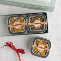 Two square metal tins of loose-leaf herbal tea displayed in a mint green gift box, with a second tin placed outside the box on a light marble surface. The tins have silver lids, round beige labels reading 'Herbal Tea,' tan bands across the tops, and a red ribbon tied in a bow next to the gift.