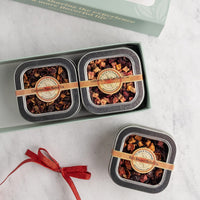 Three square metal tea tins filled with dried fruit and tea blends, arranged in and beside a pale green gift box on a light marble surface. Each tin has a tan paper band with a round label.