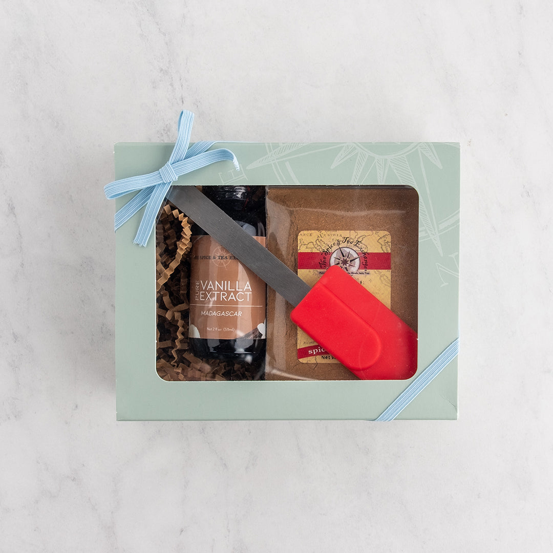 A gift box set on a white marble surface, featuring a pale green box with a light blue ribbon, showing a bottle of vanilla extract and a red silicone spatula.