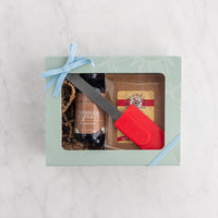 A gift box set on a white marble surface, featuring a pale green box with a light blue ribbon, showing a bottle of vanilla extract and a red silicone spatula.