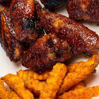 Close-up of glazed barbecue-style chicken wings piled on a white plate, with golden-orange fried tenders or strips in the foreground.