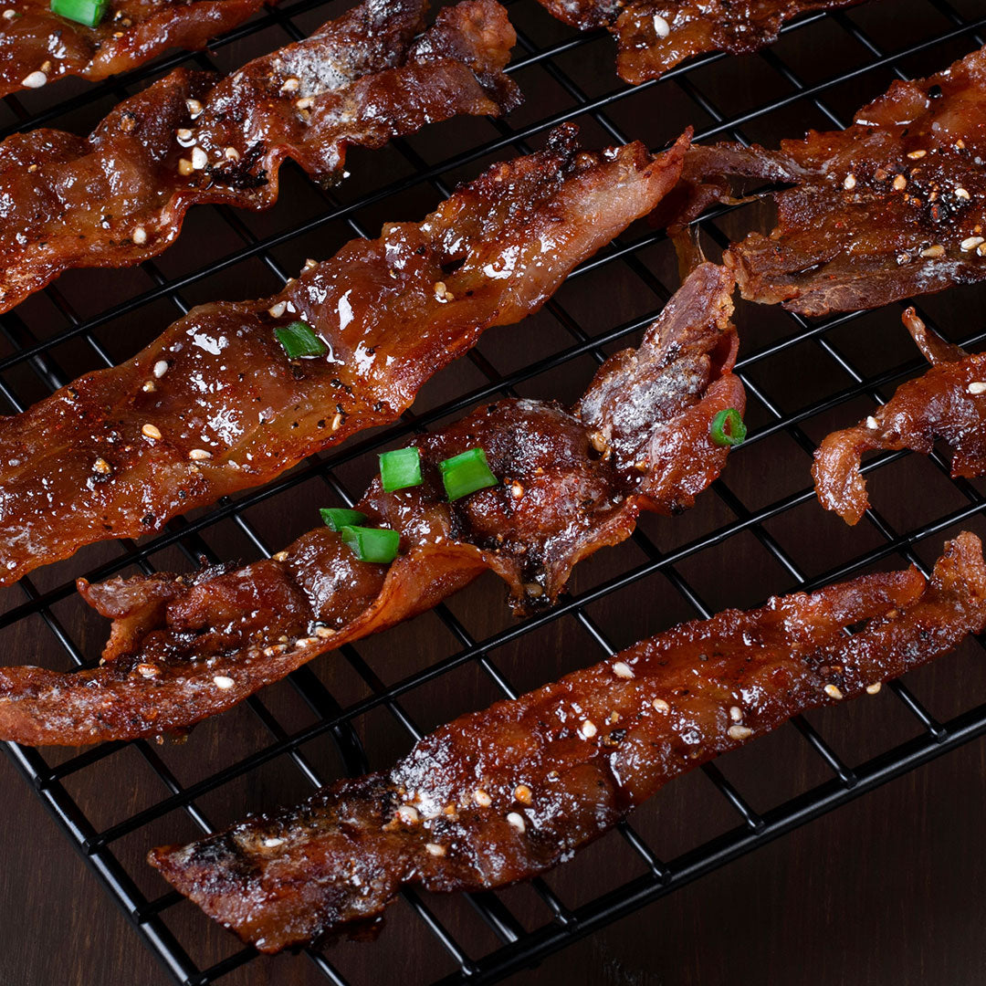Glossy, crispy glazed bacon strips sprinkled with sesame seeds and chopped green onions, arranged on a black wire cooling rack over a dark surface.