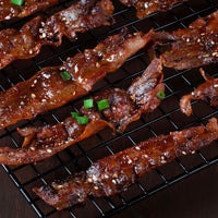 Glossy, crispy glazed bacon strips sprinkled with sesame seeds and chopped green onions, arranged on a black wire cooling rack over a dark surface.