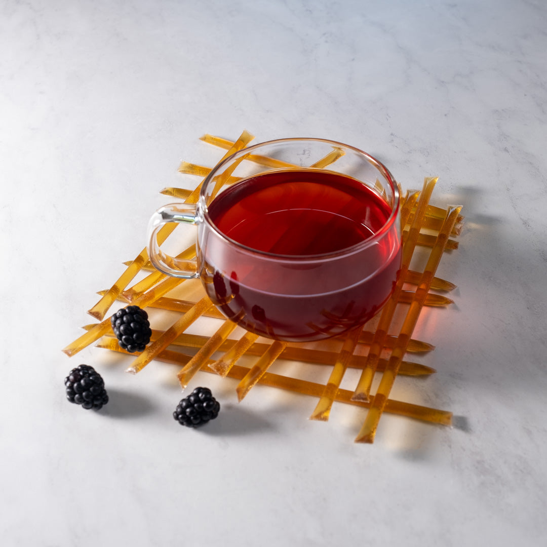 A glass cup of red herbal tea rests on a lattice mat with three blackberries and Blackberry Blossom Honey Straws nearby, all arranged on white marble—a perfect setup for a healthy snack.