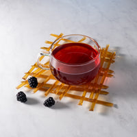 A glass cup of red herbal tea rests on a lattice mat with three blackberries and Blackberry Blossom Honey Straws nearby, all arranged on white marble—a perfect setup for a healthy snack.