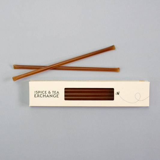 Two Bourbon Honey Straws rest on a gray surface above a white box labeled The Spice & Tea Exchange, its window displaying more Bourbon Honey Straws inside—a perfect healthy snack option.