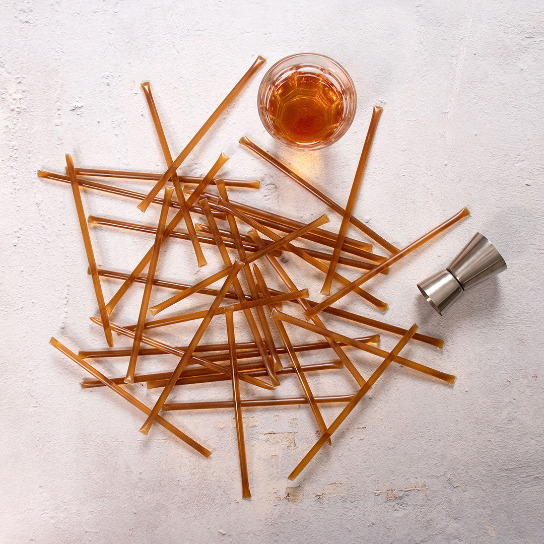 A pile of Bourbon Honey Straws is scattered on a light surface next to a glass of amber liquid and a metal jigger, echoing the golden hue of raw honey.