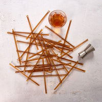 A pile of Bourbon Honey Straws is scattered on a light surface next to a glass of amber liquid and a metal jigger, echoing the golden hue of raw honey.