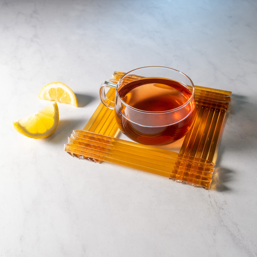 A glass cup of tea rests on a square coaster with two lemon wedges on a white marble surface—perfect for pairing with Lemon Honey Straws for a naturally sweet and healthy snack.
