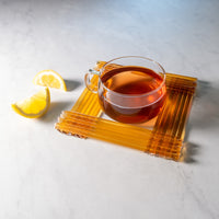 A glass cup of tea rests on a square coaster with two lemon wedges on a white marble surface—perfect for pairing with Lemon Honey Straws for a naturally sweet and healthy snack.