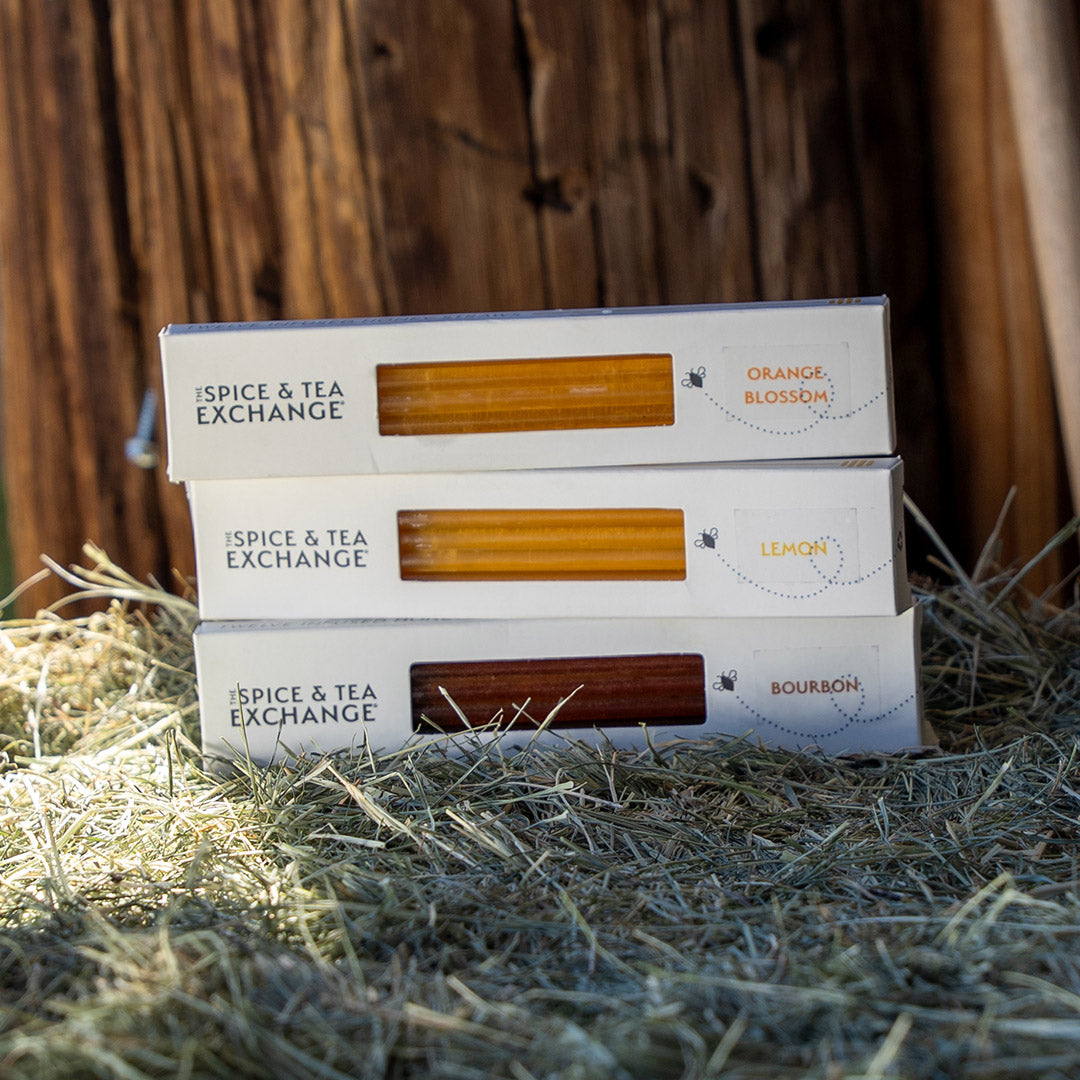 Three boxed Honey Straws from Spice & Tea Exchange stacked on hay in front of a wooden wall. The boxes are labeled Orange Blossom, Lemon, and Bourbon.