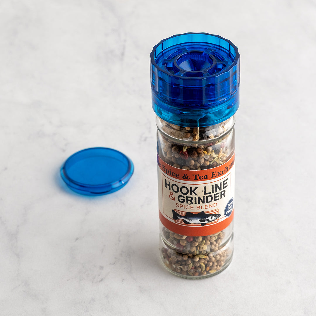A clear glass spice grinder with a translucent blue top and matching blue lid placed beside it on a light marble surface. The label reads 'Hook Line & Grinder Spice Blend' and 'The Spice & Tea Exchange,' showing ground spices inside the container.