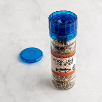A clear glass spice grinder with a translucent blue top and matching blue lid placed beside it on a light marble surface. The label reads 'Hook Line & Grinder Spice Blend' and 'The Spice & Tea Exchange,' showing ground spices inside the container.
