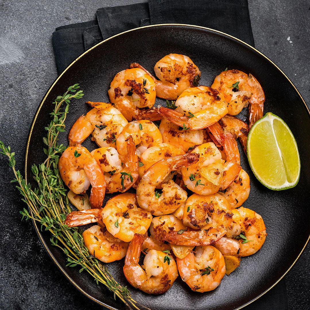 A top-down view of cooked shrimp on a black plate with a gold rim, garnished with herbs and a lime wedge, set on a dark textured surface.
