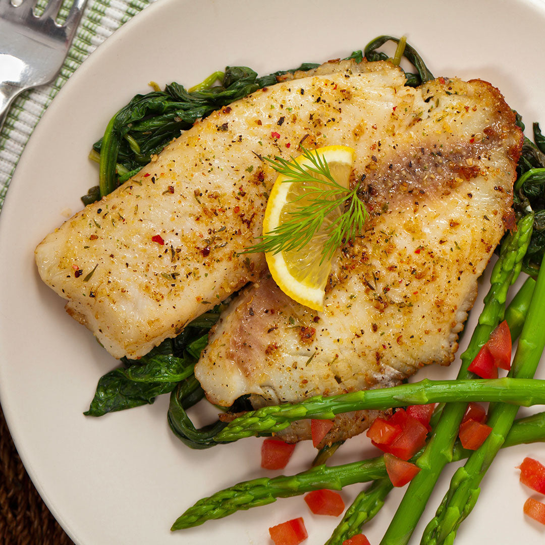 A close-up overhead shot of a plated meal featuring two seasoned fish fillets on a bed of cooked greens, served with asparagus, diced tomatoes, and a lemon slice garnish on a white plate.
