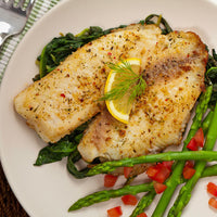 A close-up overhead shot of a plated meal featuring two seasoned fish fillets on a bed of cooked greens, served with asparagus, diced tomatoes, and a lemon slice garnish on a white plate.
