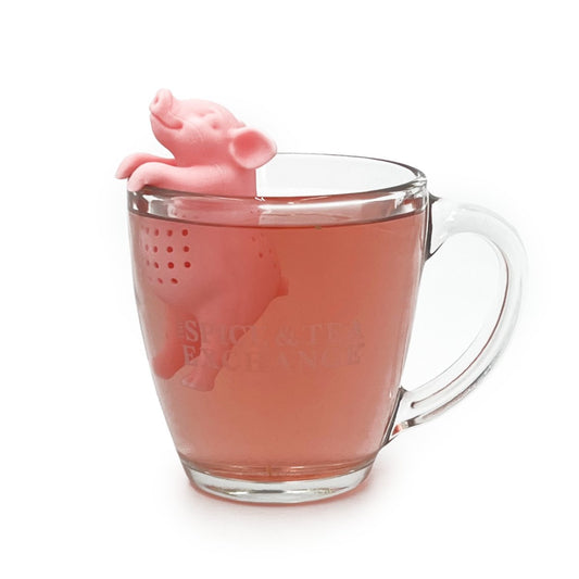 A clear glass mug holds pink tea brewed with the Hot Belly Infuser, a BPA-free silicone pig-shaped tea accessory that hooks over the rim with its head and front legs outside—ideal for making your favorite loose tea.