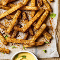 Crispy baked potato fries with a golden-brown seasoned coating, scattered on parchment paper with a small white dipping sauce bowl at the bottom.
