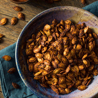 Roasted pumpkin seeds piled in a rustic blue ceramic bowl on a wooden table with scattered seeds and a blue cloth.
