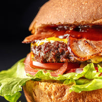 A close-up shot of a gourmet cheeseburger with a thick grilled beef patty, crispy bacon, melted yellow cheese, fresh lettuce, and sliced red onion on a dark background.
