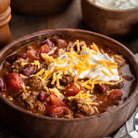 Wooden bowl of chili with toppings including sour cream and cheese on a wooden table.