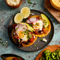 Two prepared tostadas topped with melted cheese, radishes, avocado, lime, and cilantro on a dark plate, with additional limes and food props around them on a textured blue surface.