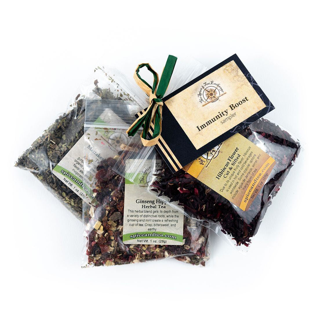 The Immunity Boost Sampler includes three clear bags of loose-leaf herbal teas—Ginseng Fig, Hibiscus Flower, and Cleansing Tea—tied with green ribbon and an “Immunity Boost Sampler” card, inspired by Marrakesh Mint Tea.