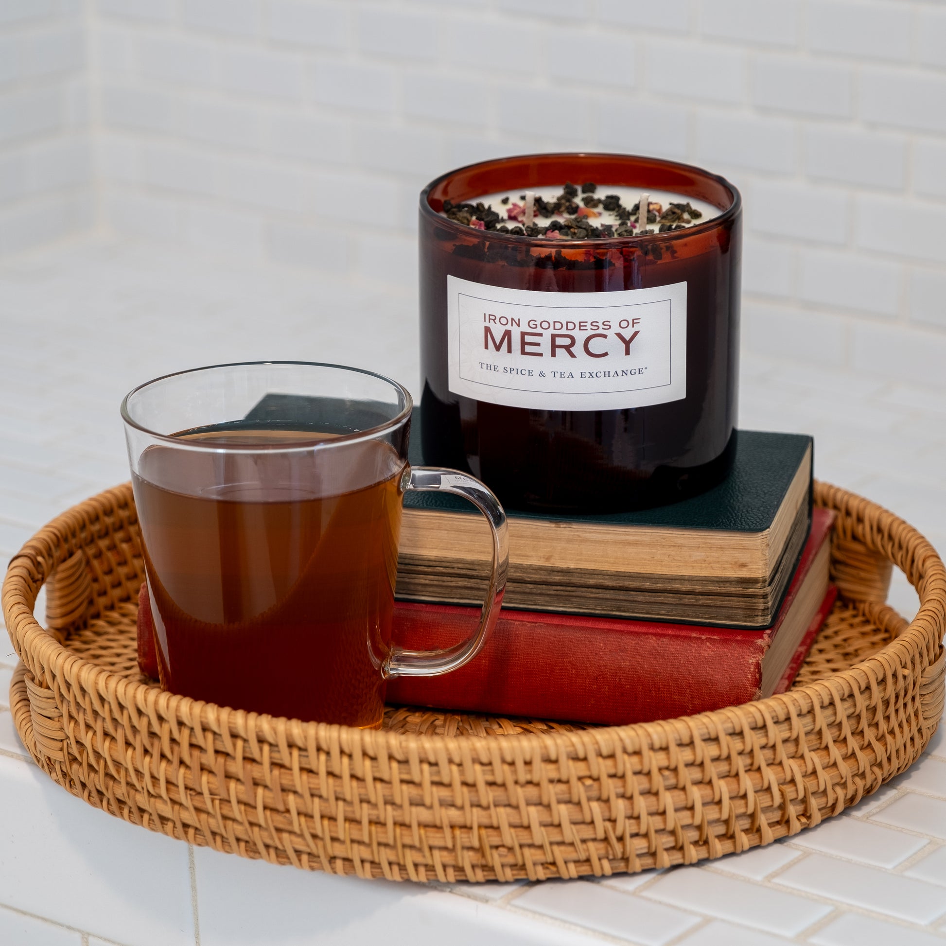 An Iron Goddess Of Mercy Candle sits on two stacked books next to a glass cup of tea on a wicker tray, with a white tiled background.