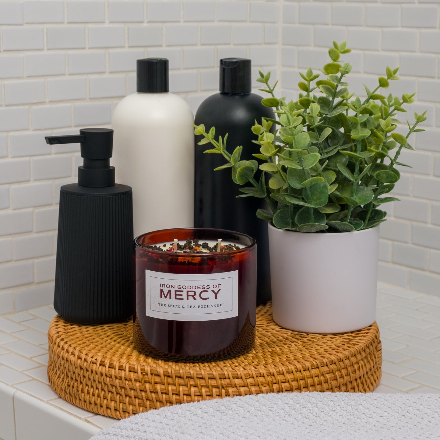 A bathroom countertop features a woven tray with a potted plant, two bottles, a black soap dispenser, and the Iron Goddess Of Mercy candle, hand poured with essential oils, all set against white tiled walls.