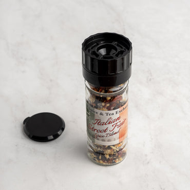 A jar of Italian Street Fair Spice Blend stands upright on a light marble surface, its black cap beside it. Inside are colorful dried spices that include smoky peppers and classic Italian seasoning.