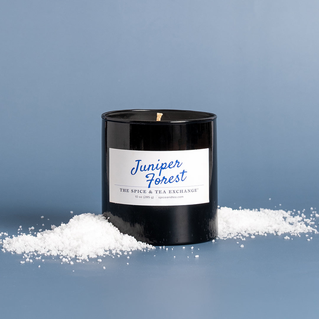 A black glass jar candle with a white label reading 'Juniper Forest' and 'The Spice & Tea Exchange' sits on a blue surface with snow scattered around the base. The background is a solid blue studio backdrop.