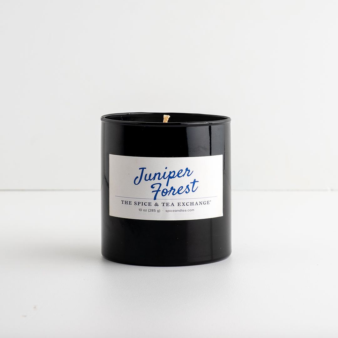 A black glass jar candle with a white label reading 'Juniper Forest' in blue script, photographed on a clean white surface against a white background.