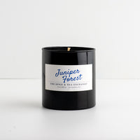 A black glass jar candle with a white label reading 'Juniper Forest' in blue script, photographed on a clean white surface against a white background.