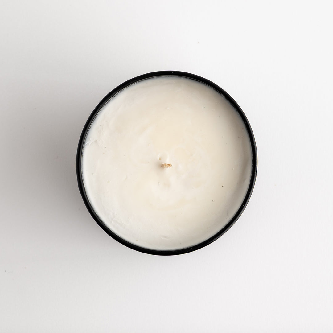 A top-down view of a single-wick candle in a round black container, filled with creamy off-white wax, set against a plain white background.