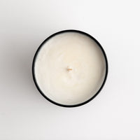 A top-down view of a single-wick candle in a round black container, filled with creamy off-white wax, set against a plain white background.