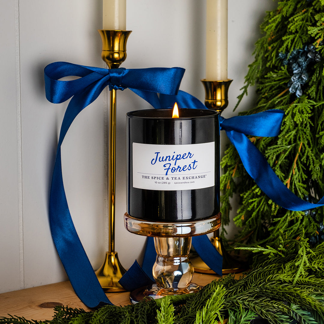 A black glass jar candle with a white label reading 'Juniper Forest' is lit and displayed on a clear glass pedestal. The candle is wrapped with a bright blue satin ribbon, styled with gold candlesticks and green pine branches against a white paneled wall.