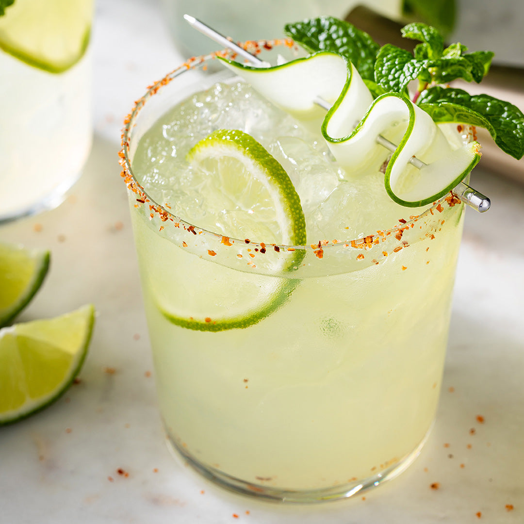 A clear margarita-style cocktail glass filled with a pale yellow drink, ice, lime wheel garnish, mint leaves, and a curled cucumber twist on a metal pick, with a salted rim and lime slices on a light marble surface.