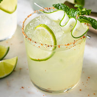 A clear margarita-style cocktail glass filled with a pale yellow drink, ice, lime wheel garnish, mint leaves, and a curled cucumber twist on a metal pick, with a salted rim and lime slices on a light marble surface.