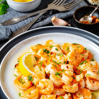 A close-up shot of a plate of cooked shrimp prawns garnished with chopped green chives, garlic, and a lemon wedge, served on a white plate with a dark rim.