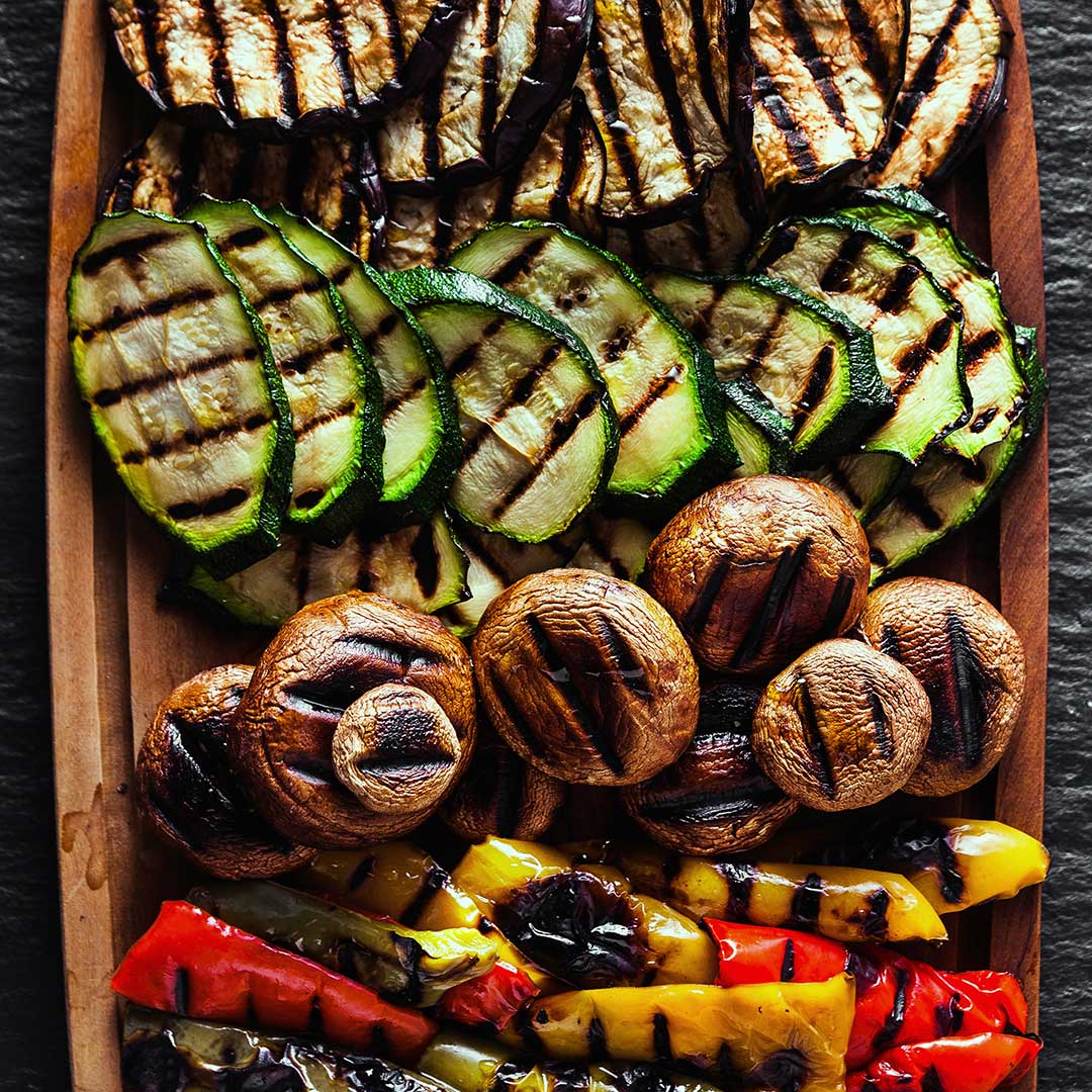 Grilled vegetables on a wooden serving board, including sliced zucchini, mushrooms, red and yellow bell peppers, and grilled eggplant, photographed from above on a dark slate background.