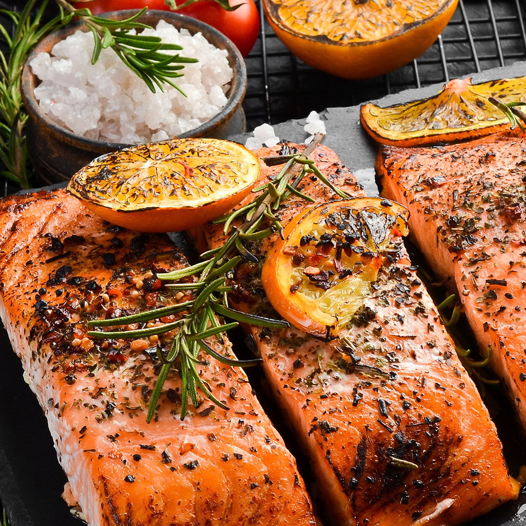Three grilled salmon fillets on a dark plate, garnished with roasted orange slices, rosemary sprigs, and black pepper, with bowls of salt and orange halves in the background.