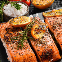 Three grilled salmon fillets on a dark plate, garnished with roasted orange slices, rosemary sprigs, and black pepper, with bowls of salt and orange halves in the background.