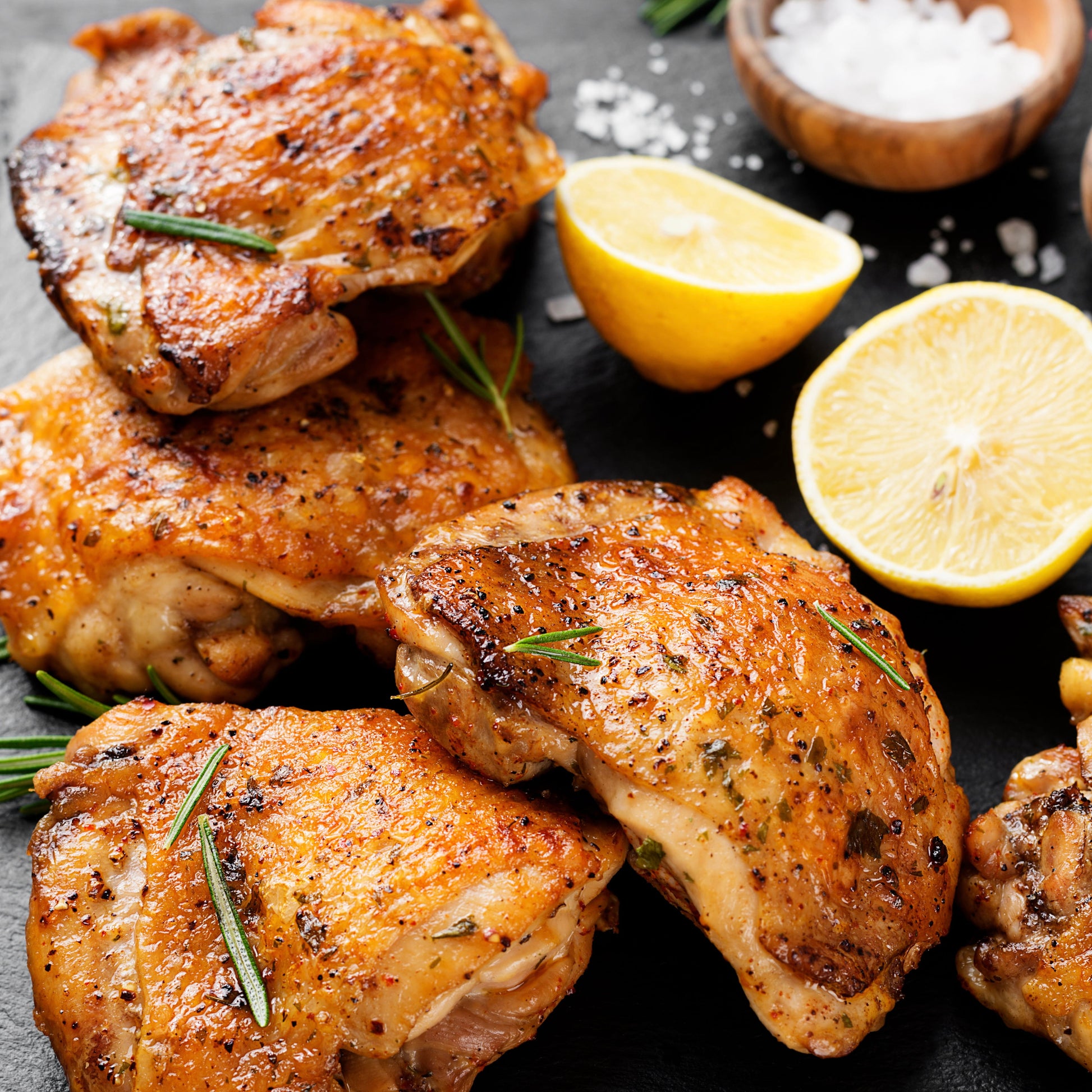 Crispy roasted chicken thighs garnished with rosemary and herbs, arranged on a dark surface with lemon halves, coarse salt, and a small bowl of white seasoning.
