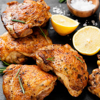 Crispy roasted chicken thighs garnished with rosemary and herbs, arranged on a dark surface with lemon halves, coarse salt, and a small bowl of white seasoning.
