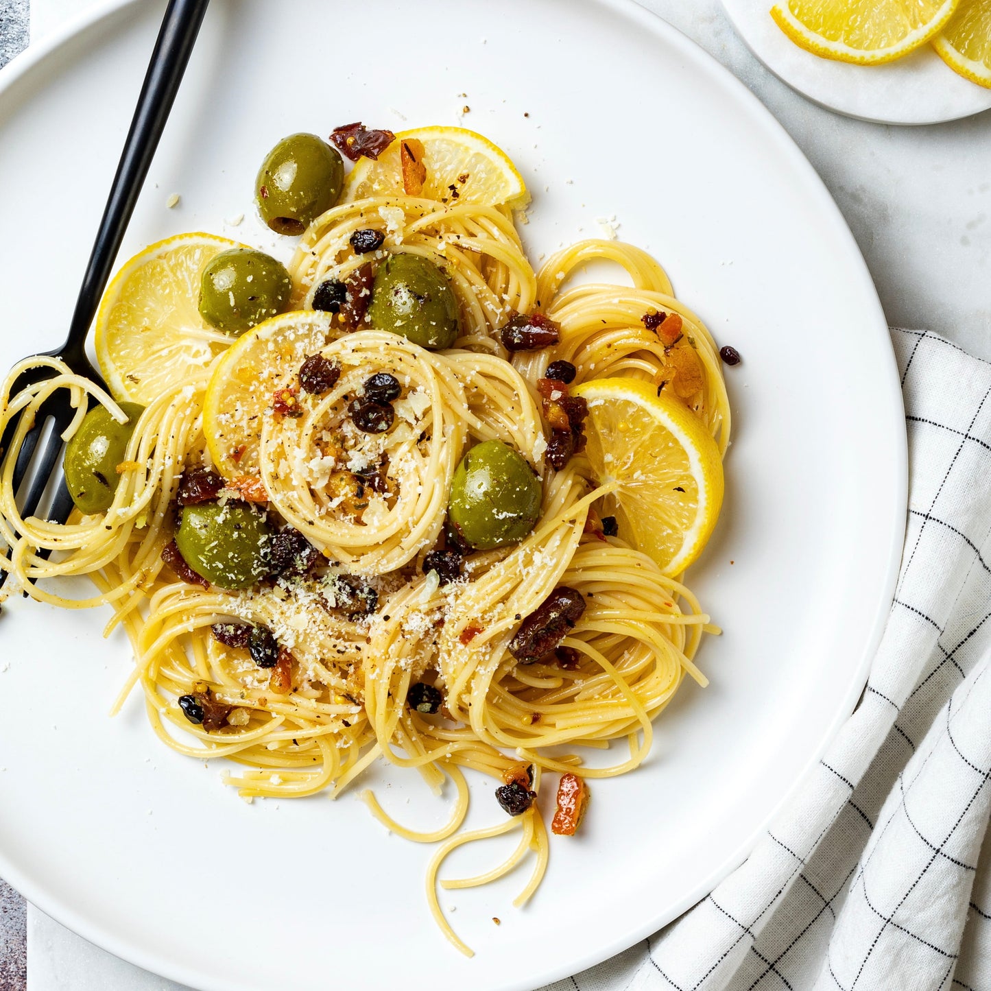 A plated pasta dish on a white plate with lemon slices, green olives, dried fruit pieces, and a black fork, photographed on a light marble-like surface with a folded checkered cloth.