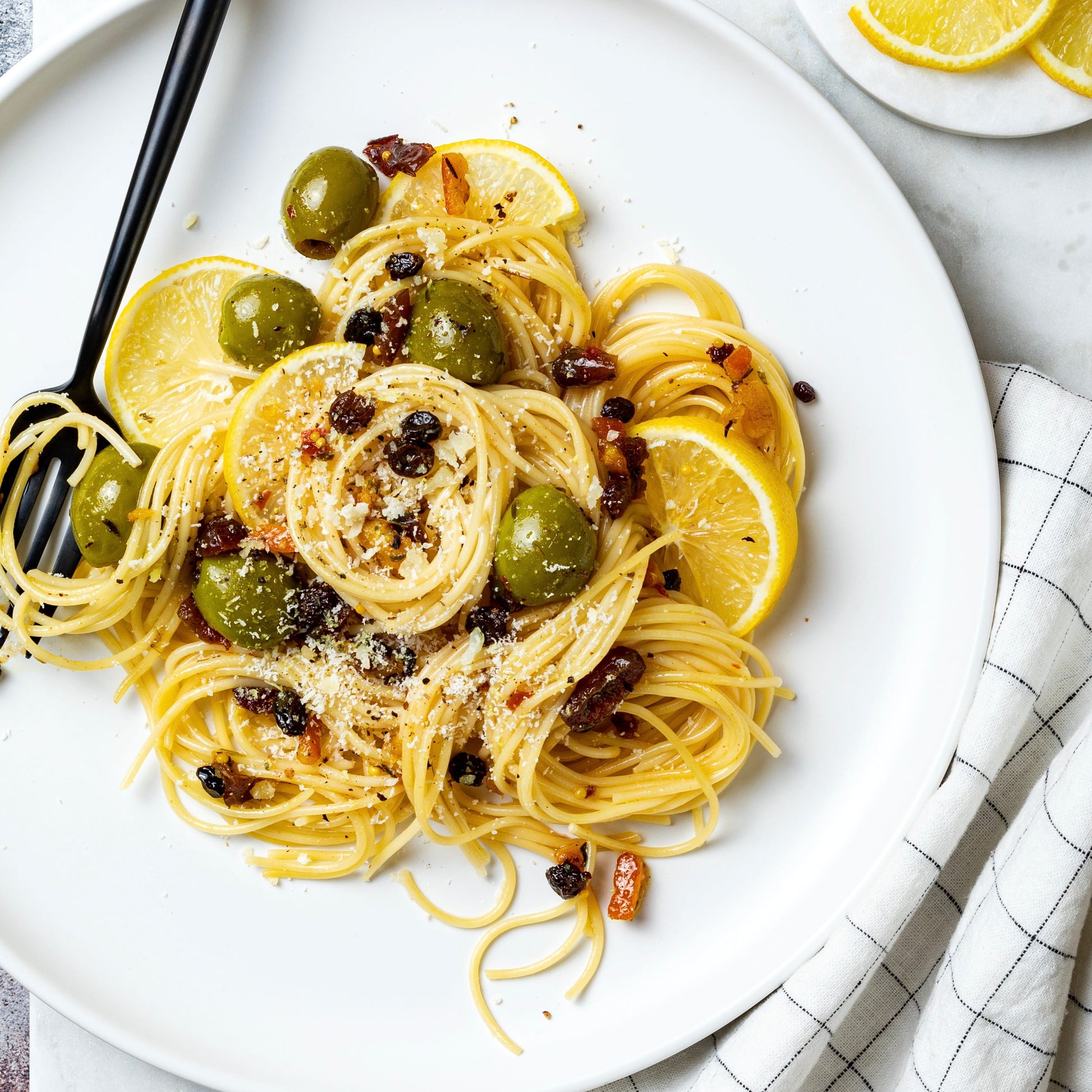 A plated pasta dish on a white plate with lemon slices, green olives, dried fruit pieces, and a black fork, photographed on a light marble-like surface with a folded checkered cloth.