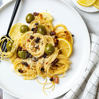 A plated pasta dish on a white plate with lemon slices, green olives, dried fruit pieces, and a black fork, photographed on a light marble-like surface with a folded checkered cloth.