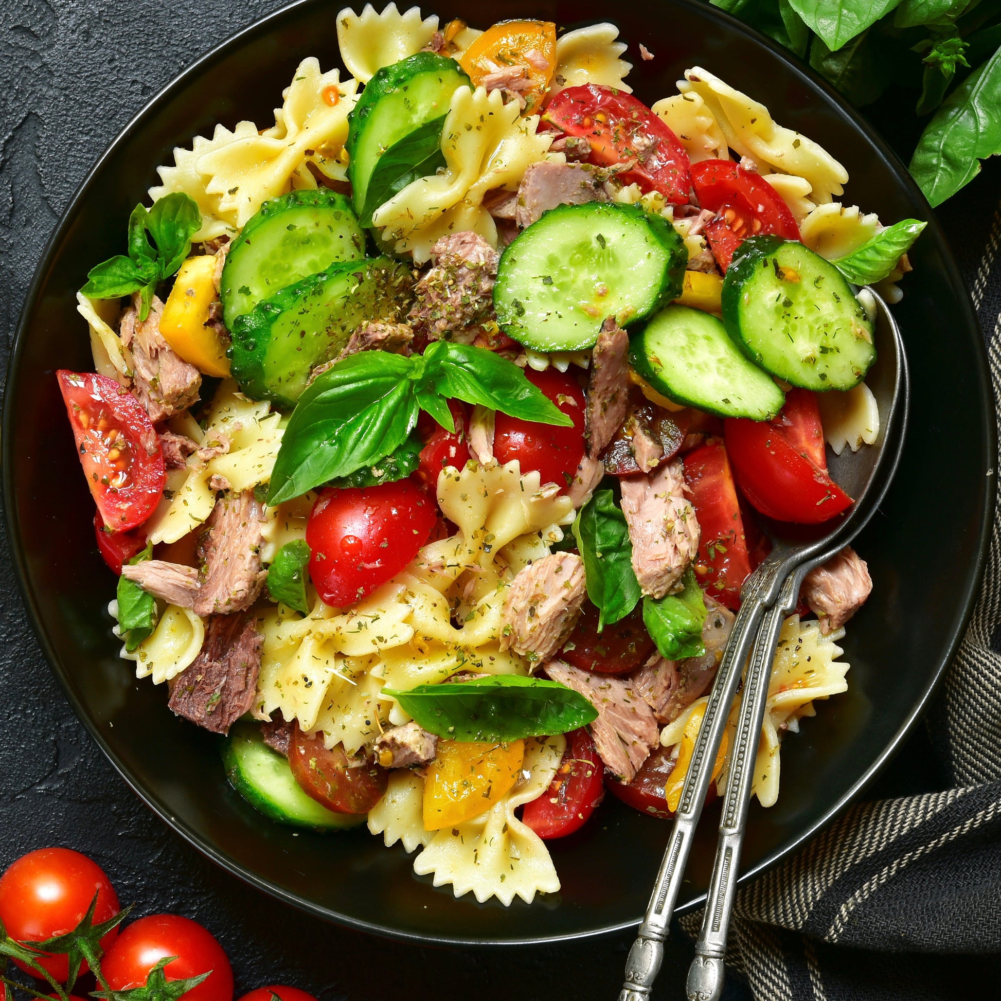 A top-down view of a fresh pasta salad in a black bowl, featuring bowtie pasta, sliced cucumbers, tomatoes, tuna, and fresh basil leaves, with silver serving spoons resting on the side.