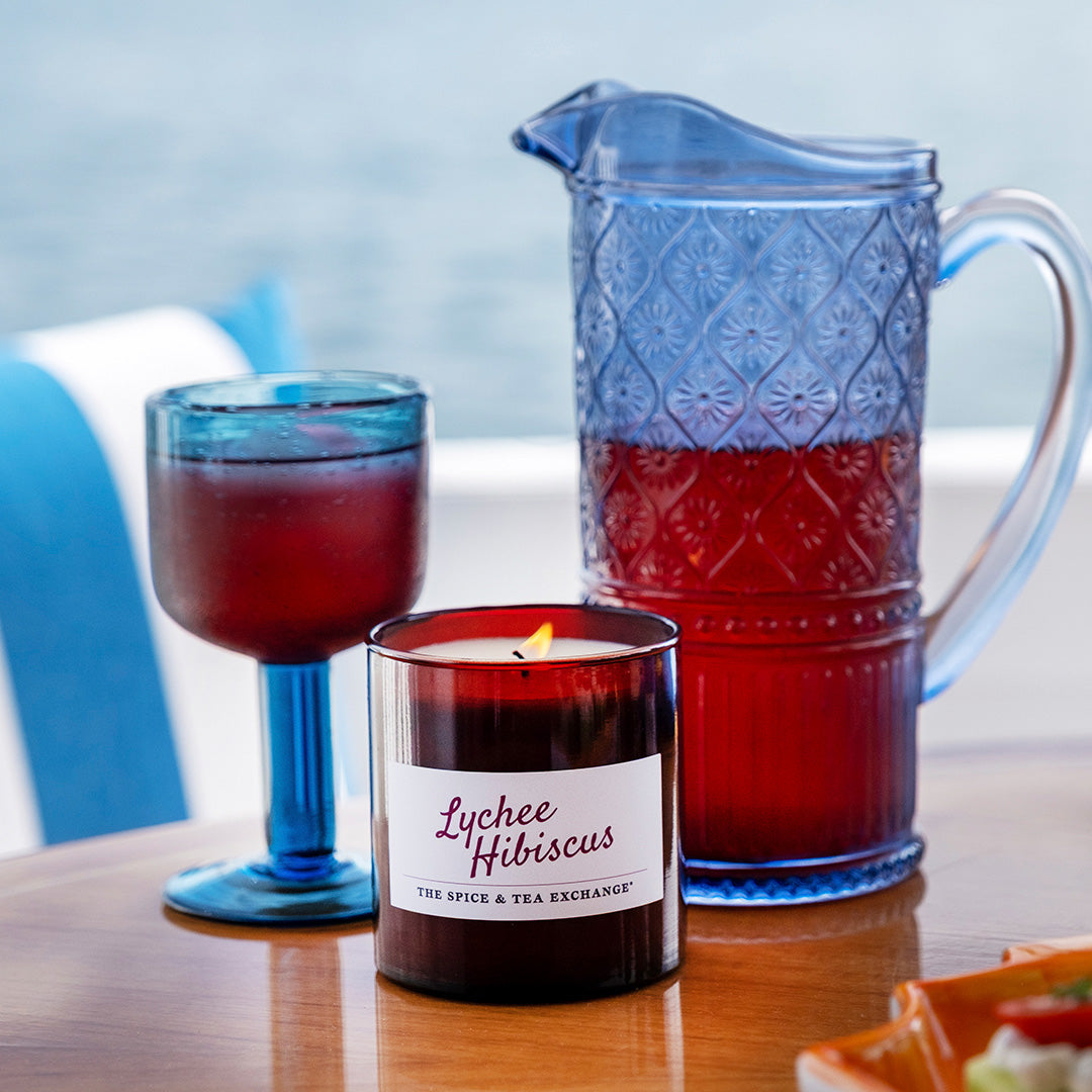 Candle labeled 'Lychee Hibiscus' next to a glass and pitcher on a table with a blurred outdoor background.